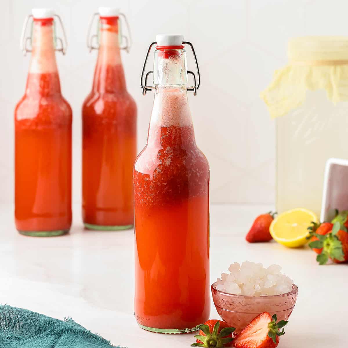 A bottle of strawberry water kefir with a flip top lid, surrounded by a small bowl of water kefir grains, fresh strawberries, lemons, and more bottles of strawberry kefir. 