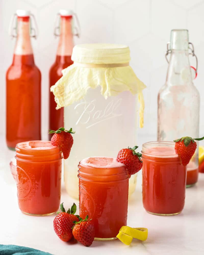 Several jars and bottles of fermented strawberry kefir water, with a jar of cheesecloth covered regular kefir water.