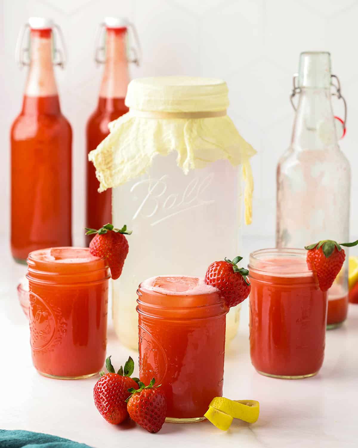 Several jars and bottles of fermented strawberry kefir water, with a jar of cheesecloth covered regular kefir water. 