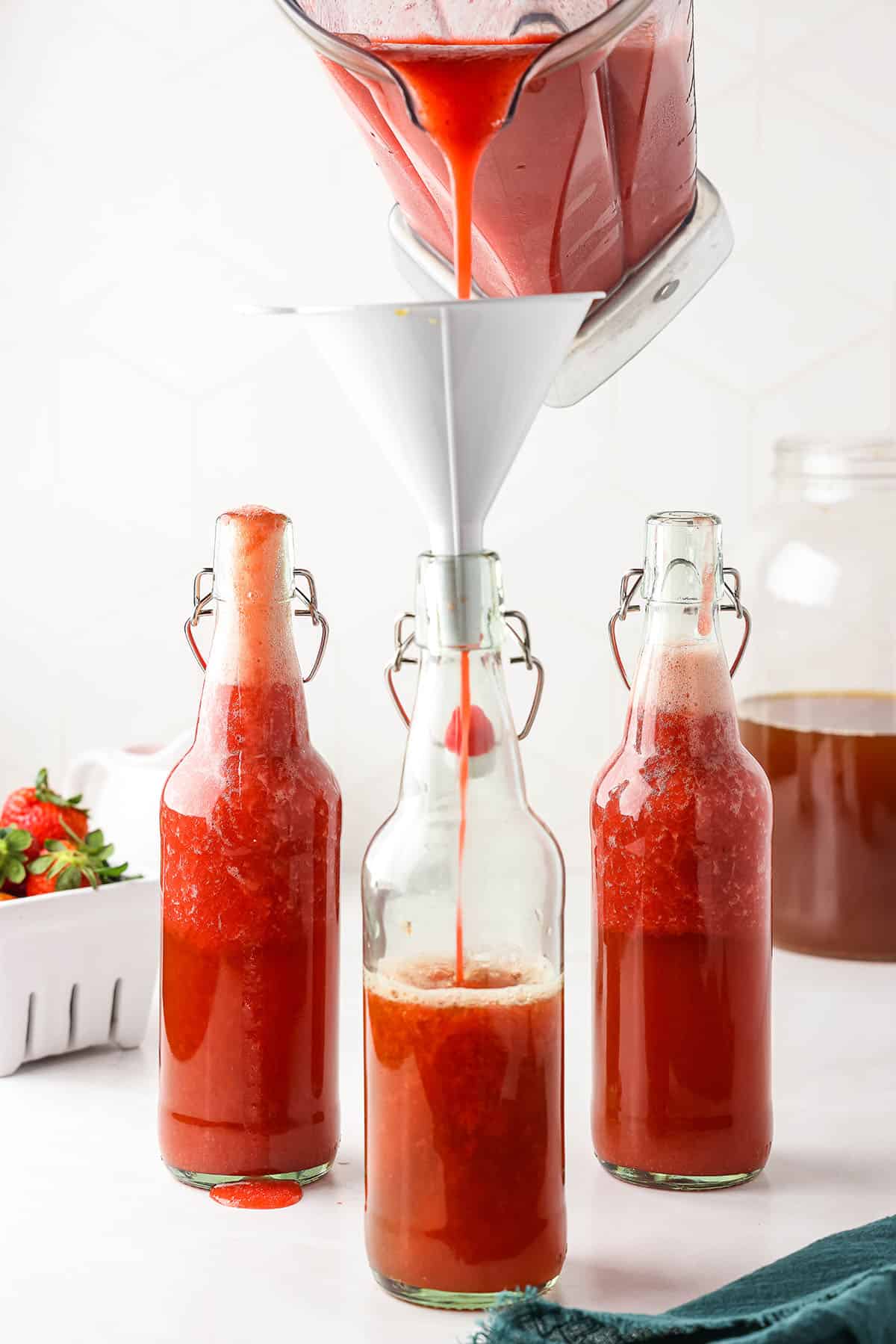 Flip top bottles with strawberry puree and kombucha in them, one with a funnel on top and puree pouring in.
