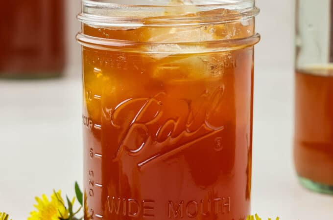 A glass jar of dandelion kombucha with ice, surrounded by fresh dandelion flowers.