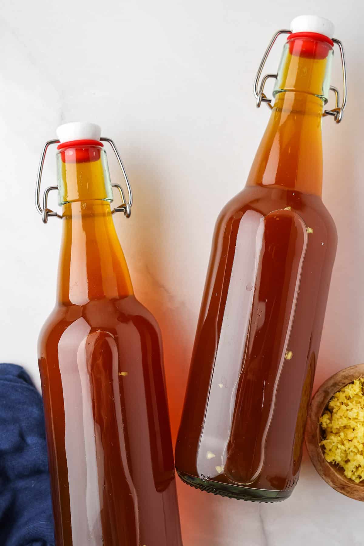 2 Bottles of ginger kombucha laying on their sides, with a bowl of fresh minced ginger next to it. 