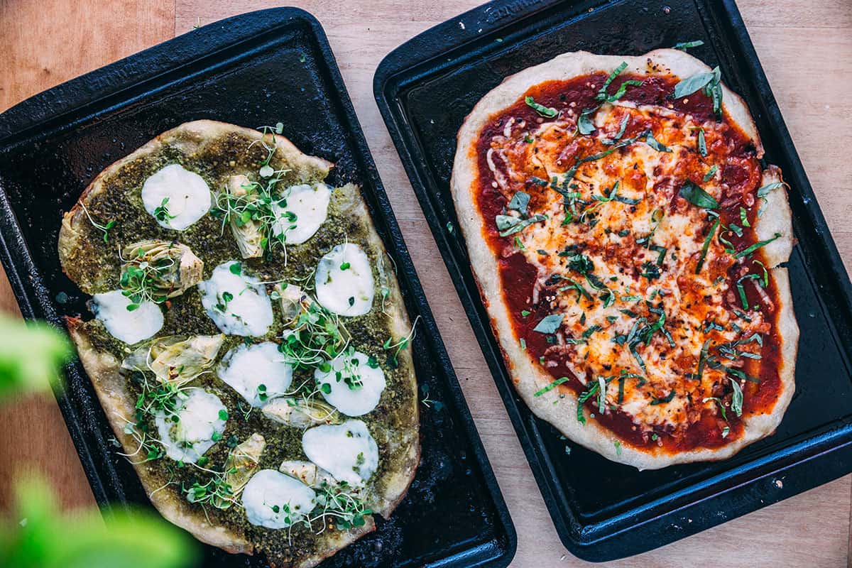 Two baking sheets with sourdough pizzas with different toppings on them. 