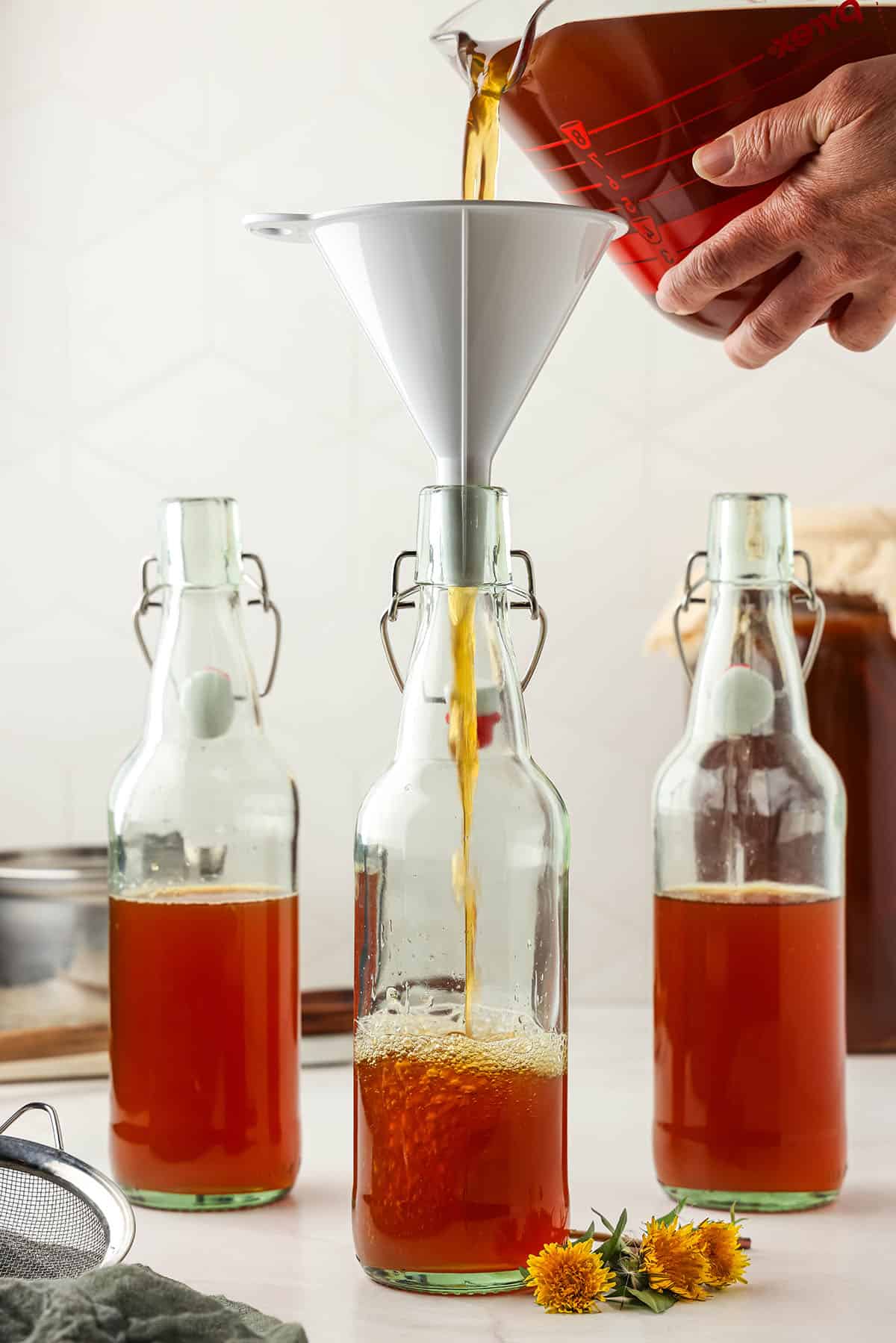 Several bottles filled halfway with basic kombucha, and one pouring in with a funnel. 