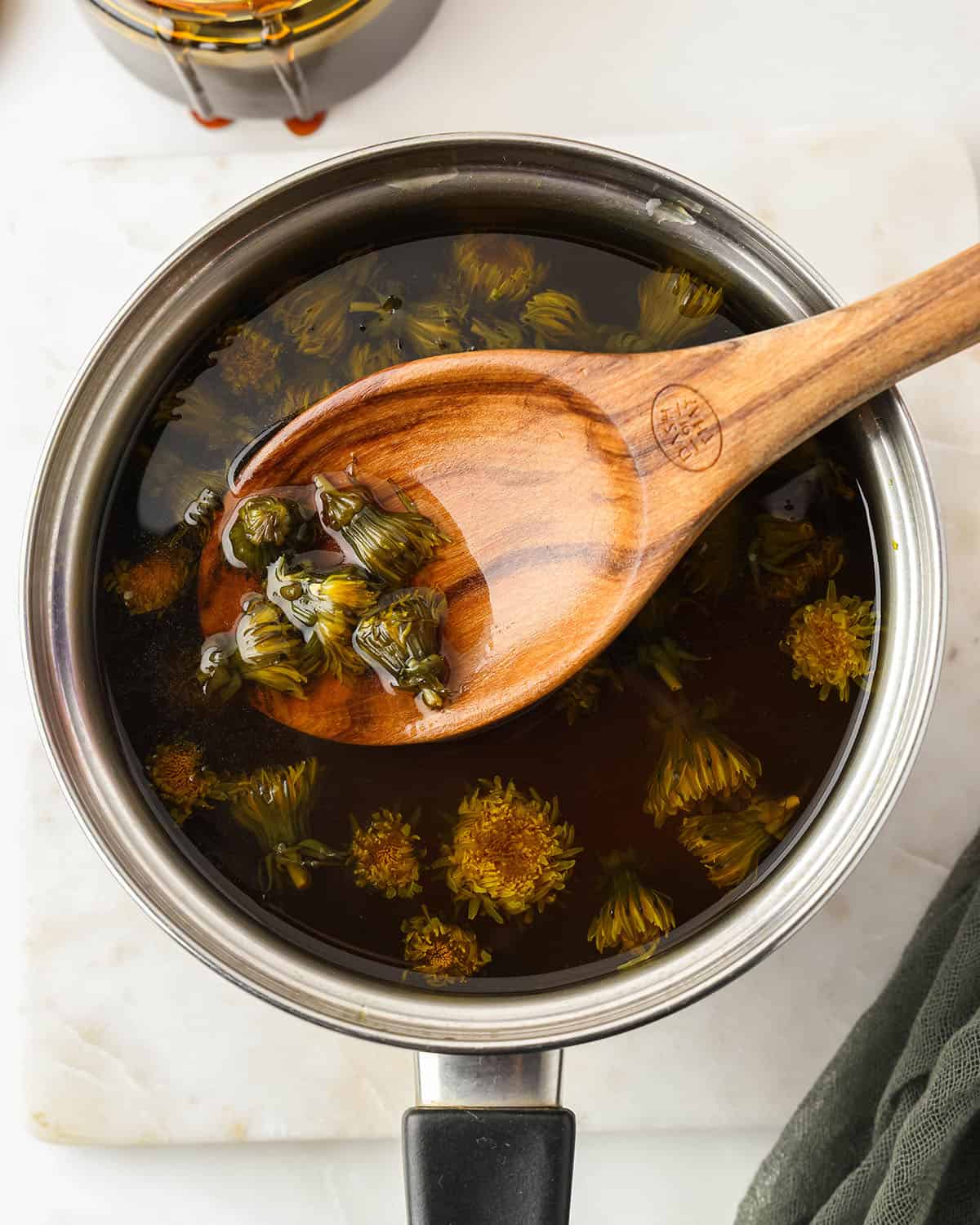 Dandelions steeping in hot water in a pot for tea with a wooden spoon lifting the blossoms out. 