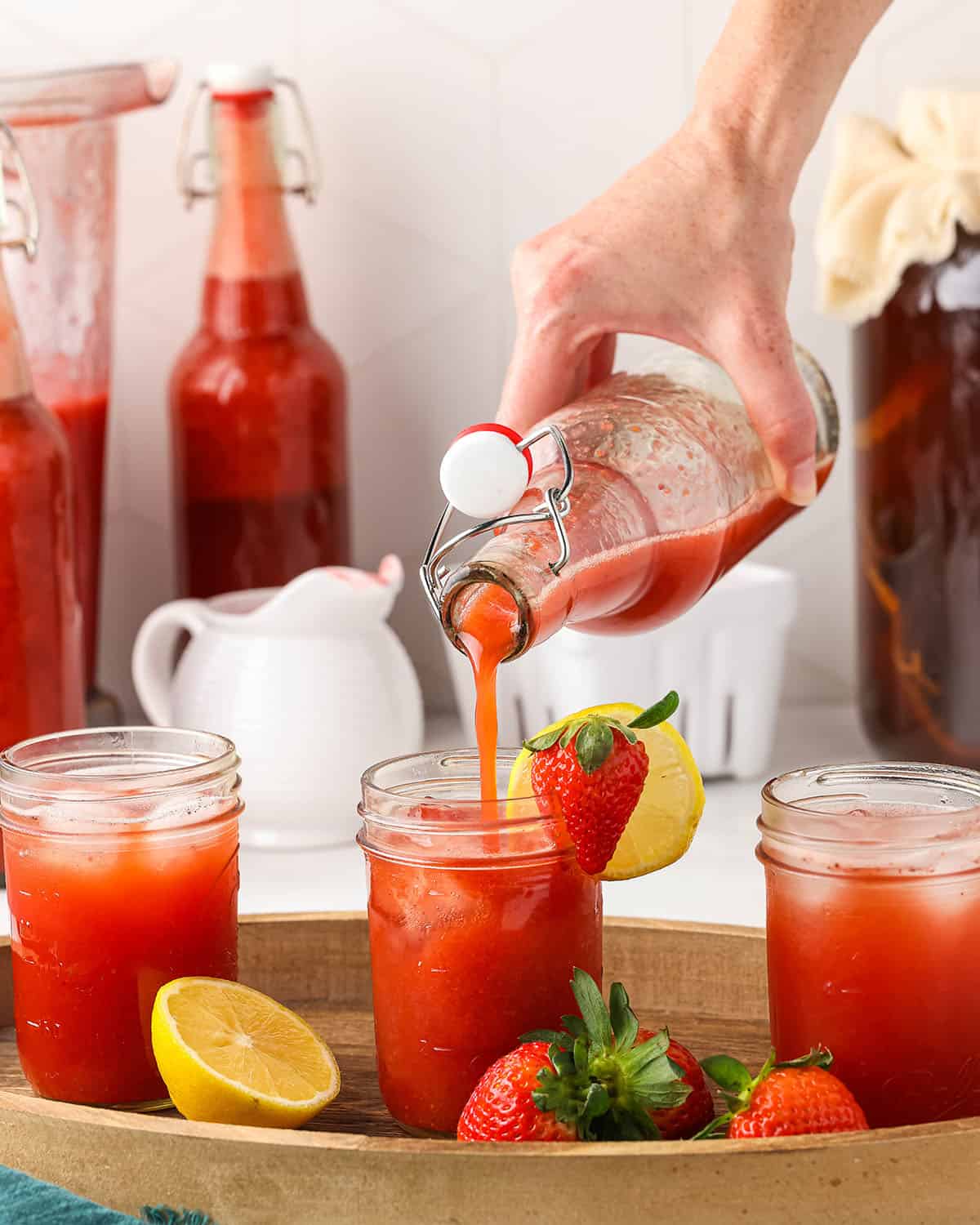 A bottle of red strawberry kombucha pouring into a glass, surrounded by other glasses of strawberry kombucha, and fresh fruit. 