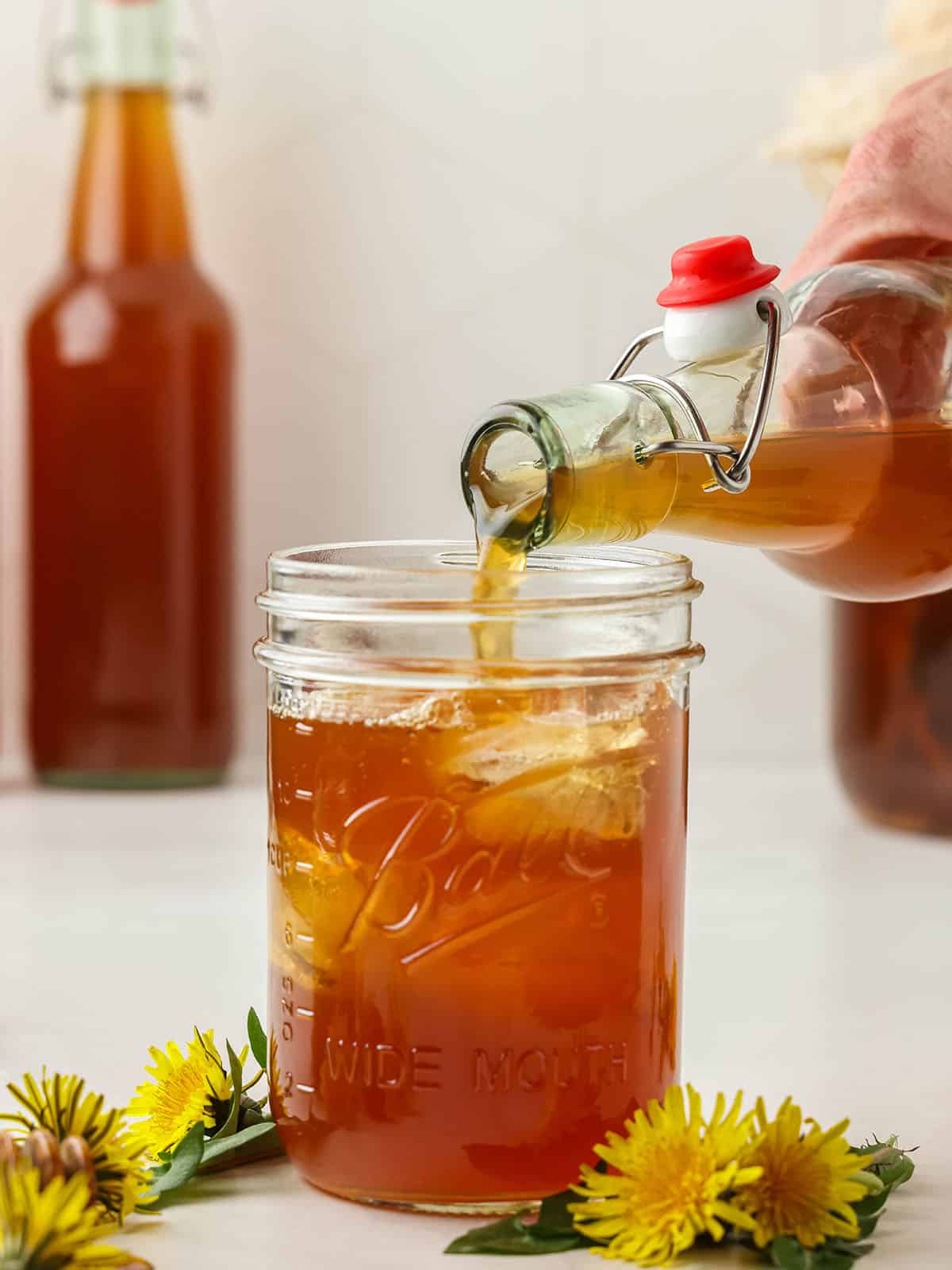 Dandelion kombucha pouring into a glass with ice, with fresh dandelions surrounding. 