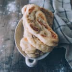 A stack of naan bread in a bowl on a wood surface with flour on it and a kitchen cloth surrounding.