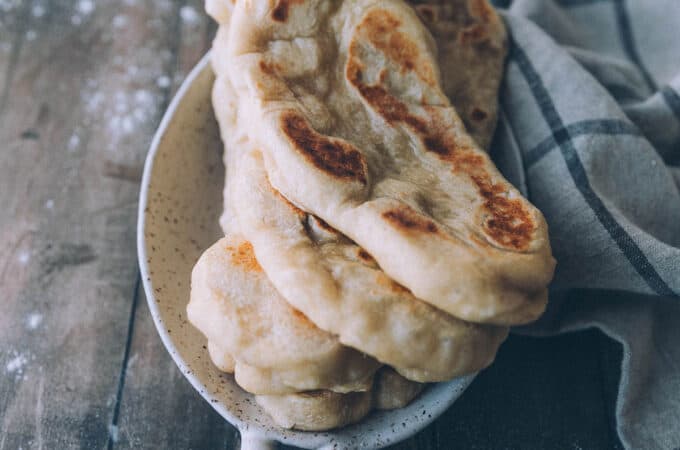 A stack of naan bread in a bowl on a wood surface with flour on it and a kitchen cloth surrounding.