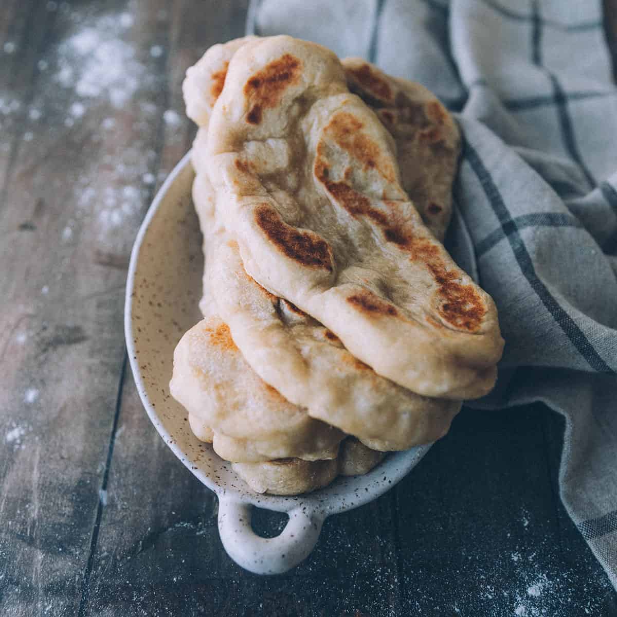 A stack of naan bread in a bowl on a wood surface with flour on it and a kitchen cloth surrounding. 