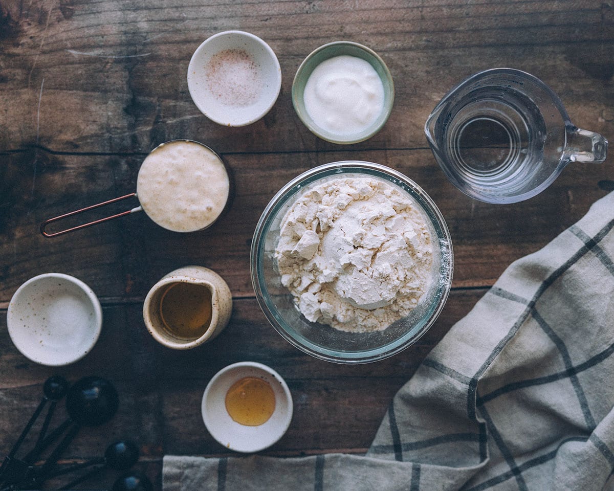 Sourdough naan ingredients in small bowls on a wood surface, top view. 