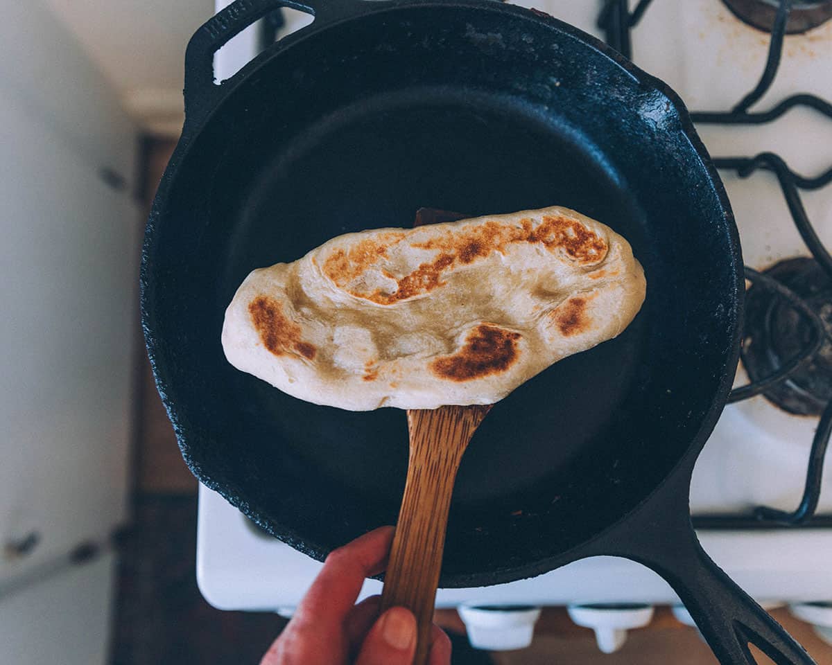 Sourdough naan being flipped while being cooked in a cast iron pan. 