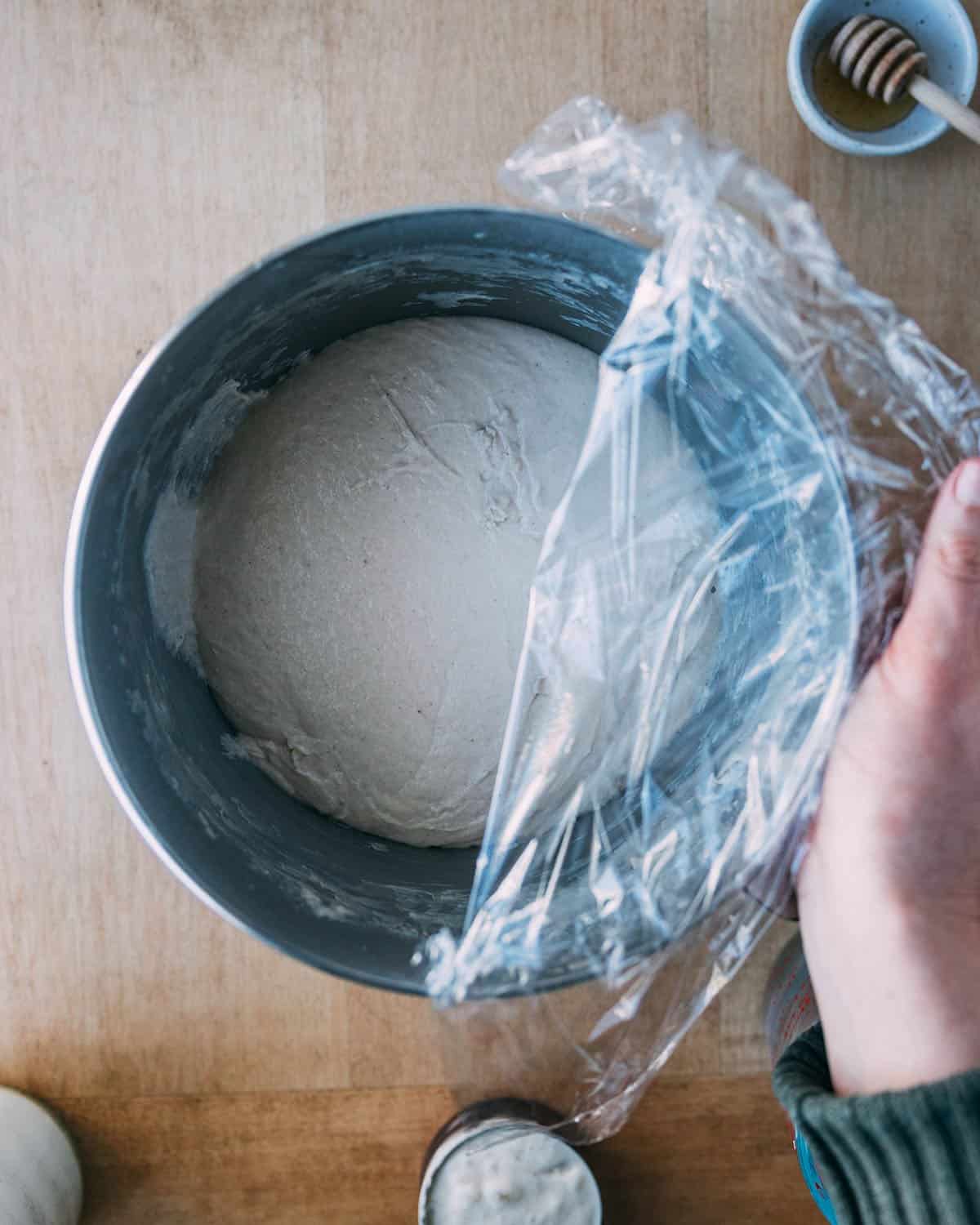 Sourdough pizza dough after rising with the plastic wrap half off the bowl, top view. 