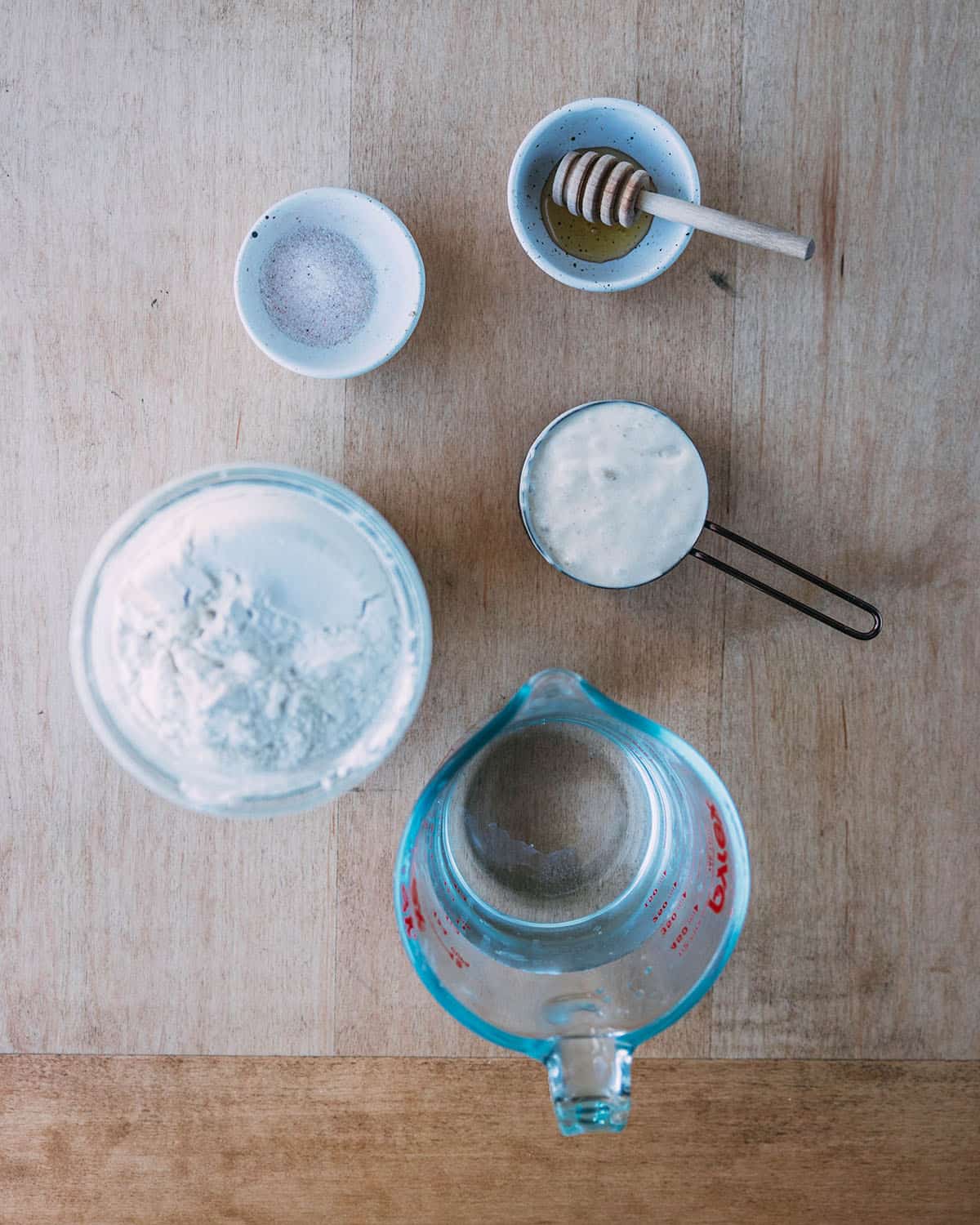 Sourdough pizza dough ingredients in small bowls and measuring cups on a light wood surface, top view. 