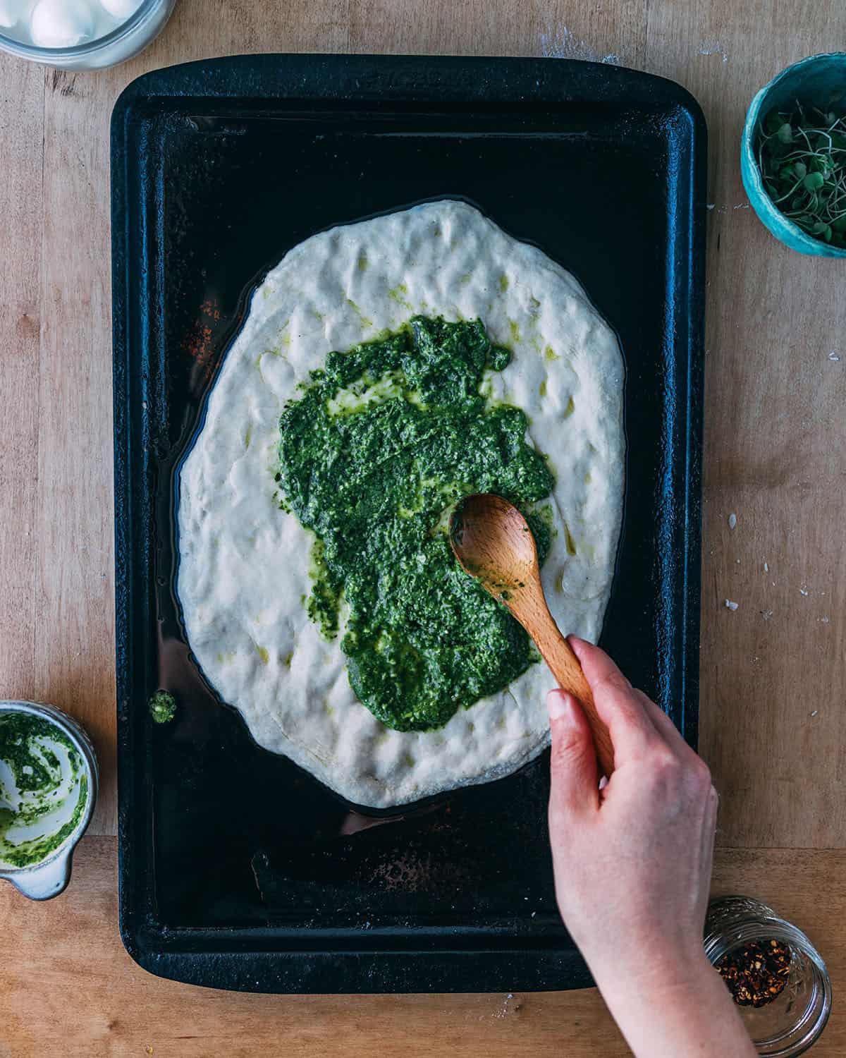 Sourdough pizza dough on a sheet pan with pesto being spread onto it. 