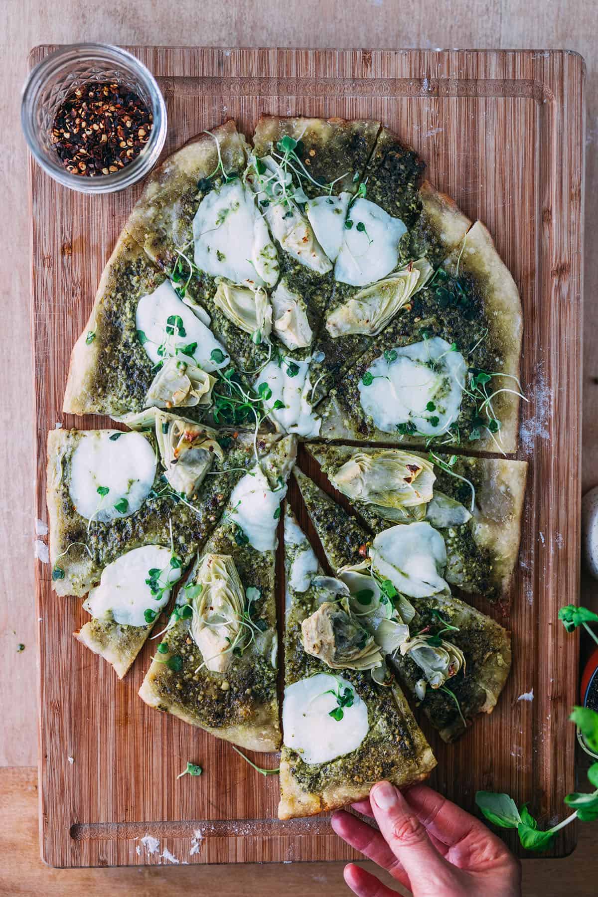A sourdough pizza with pesto, artichoke hearts, and mozzarella sliced on a wood cutting board.  