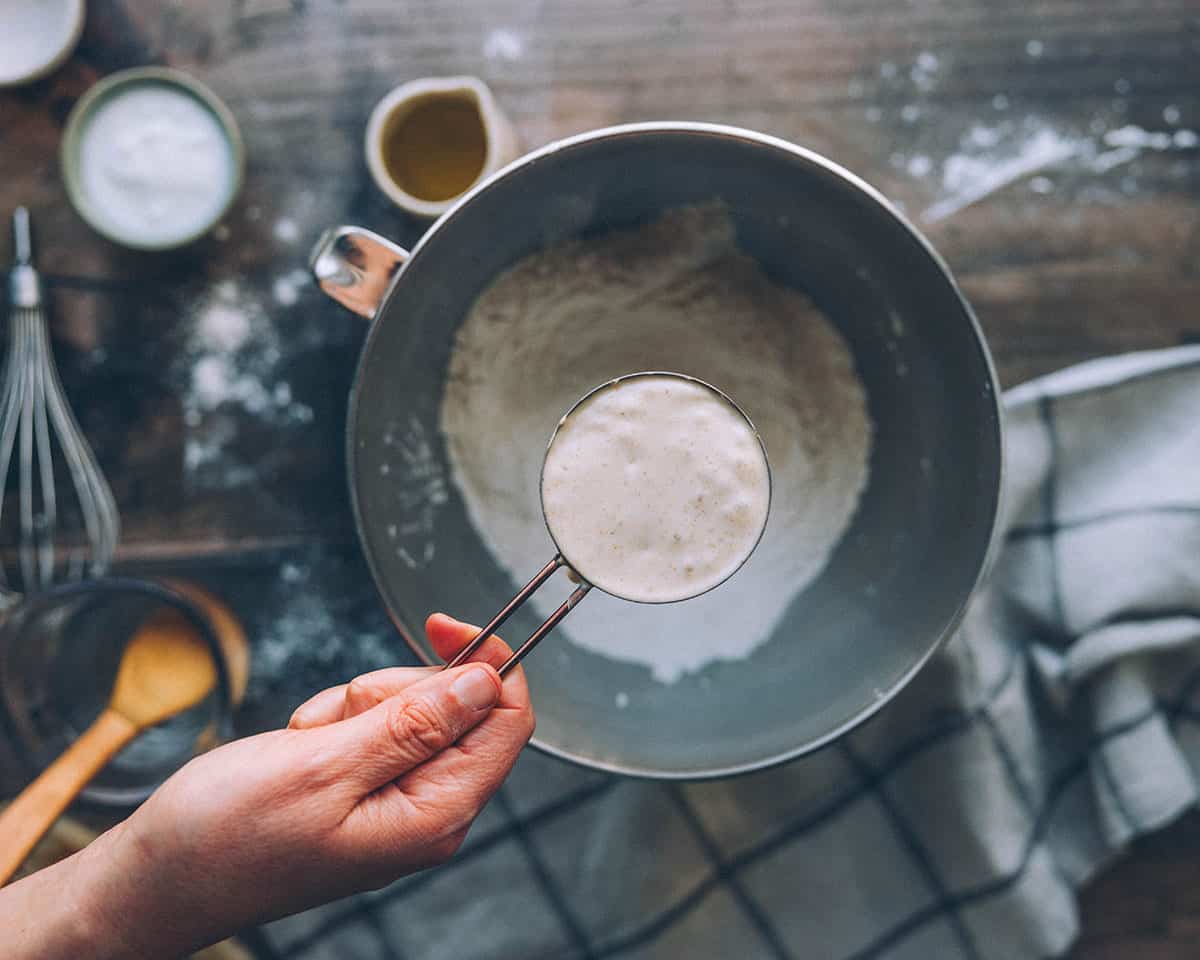 A hand pouring a measuring cup of sourdough starter into the dry ingredients. 