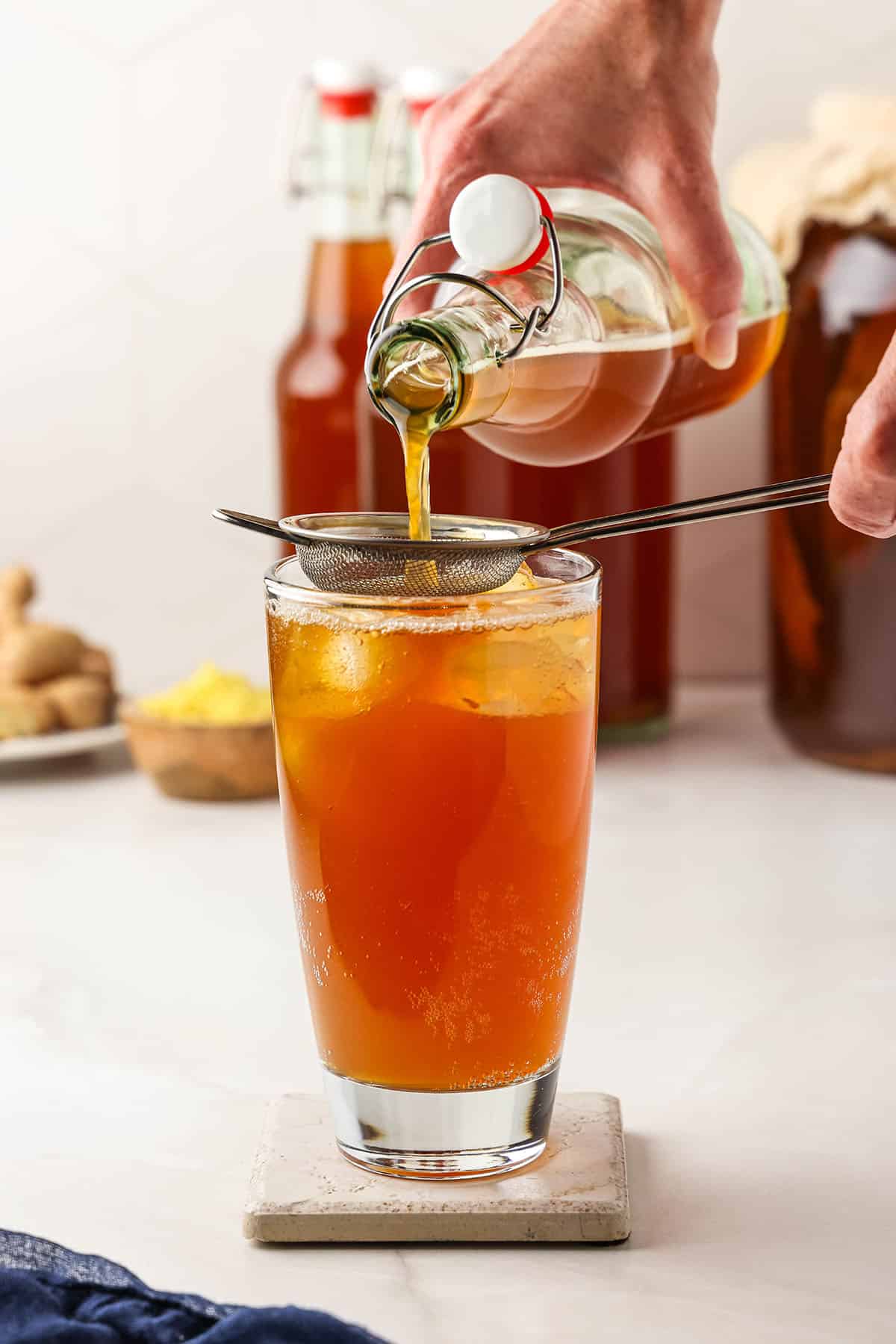 A bottle of ginger kombucha pouring through a strainer to reserve the ginger bits, into a tall glass with ice. 