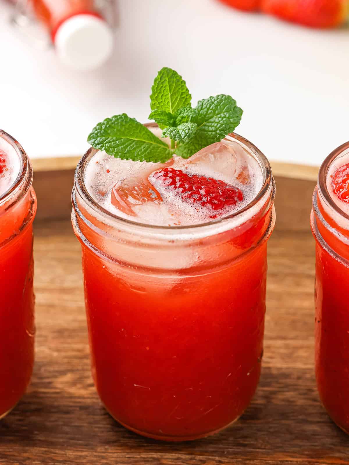 A glass with strawberry kombucha in it, with ice and some mint leaves, on a wood surface. 