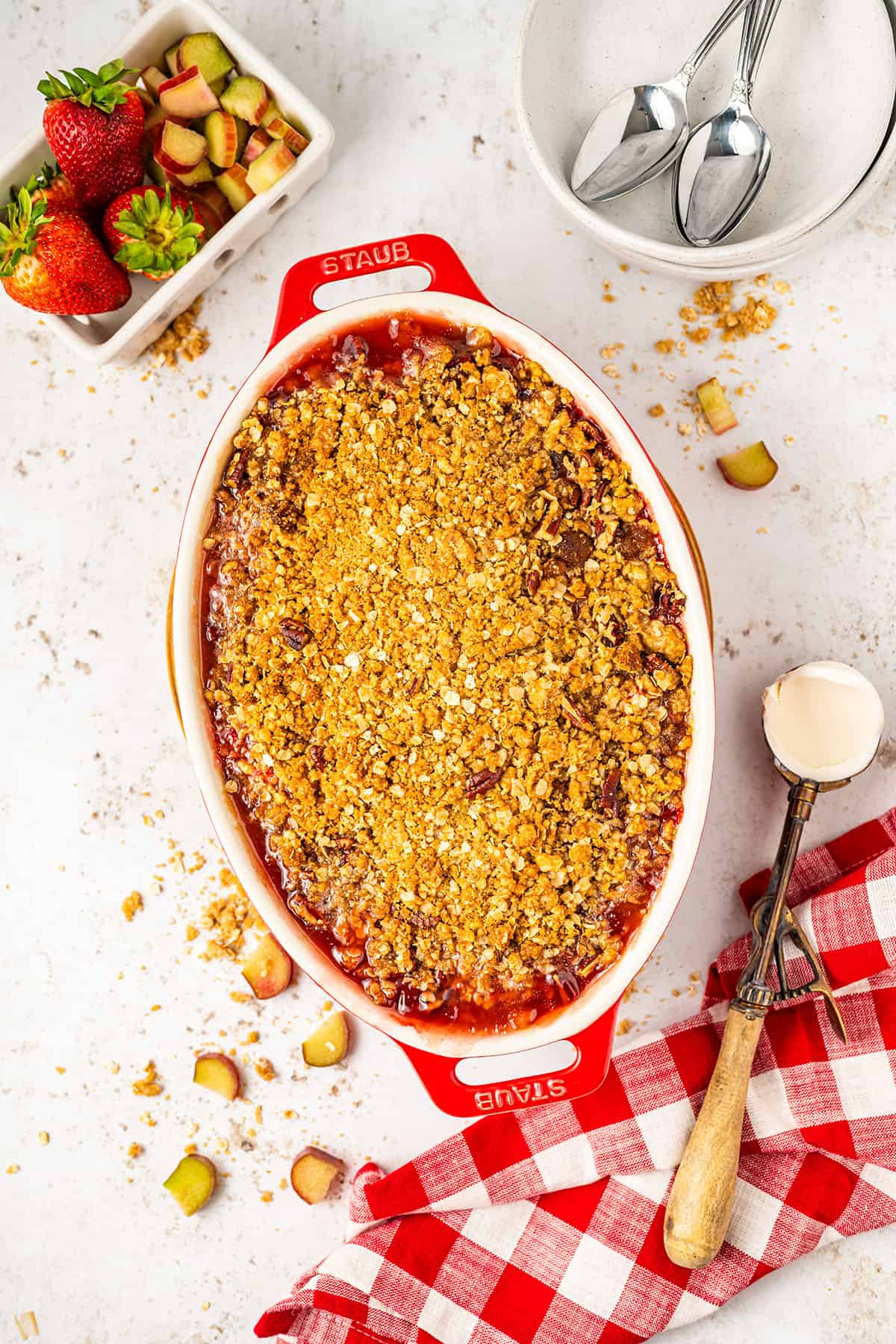 Baked strawberry rhubarb crisp in a baking dish, on a white counter surrounded by a red and white checked cloth, fresh strawberries, and an ice cream scoop. 