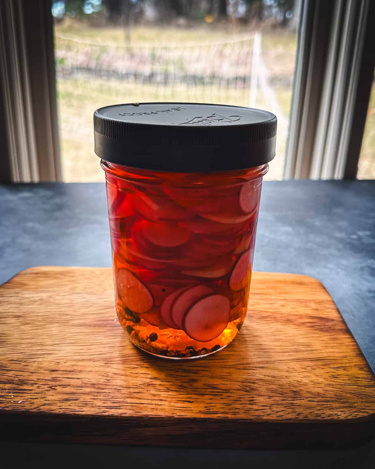 A jar of pickled radishes with a lid on a cutting board in front of a window.