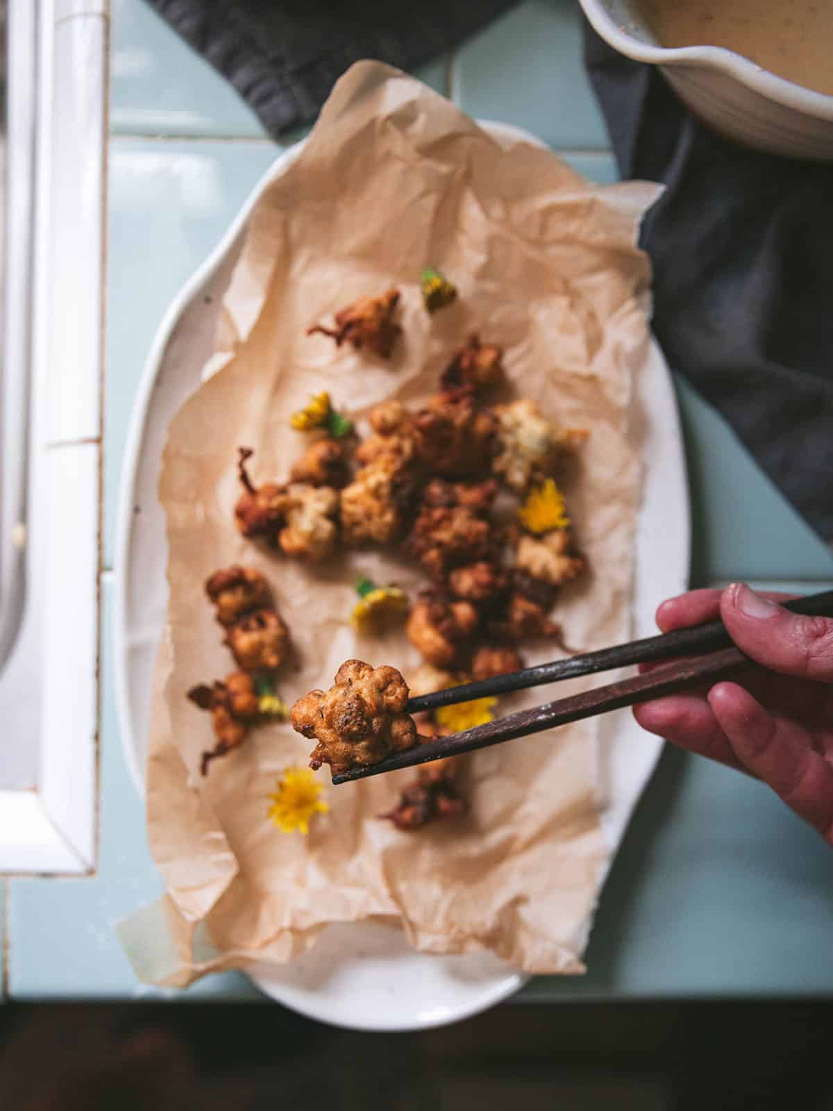 Dandelion fritters on a natural napkin on an oval white plate, with chopsticks lifting one up. 