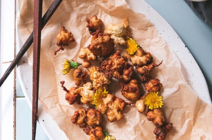 An oval plate with a natural napkin holding fried dandelion fritters, with chopsticks across the top of the plate.