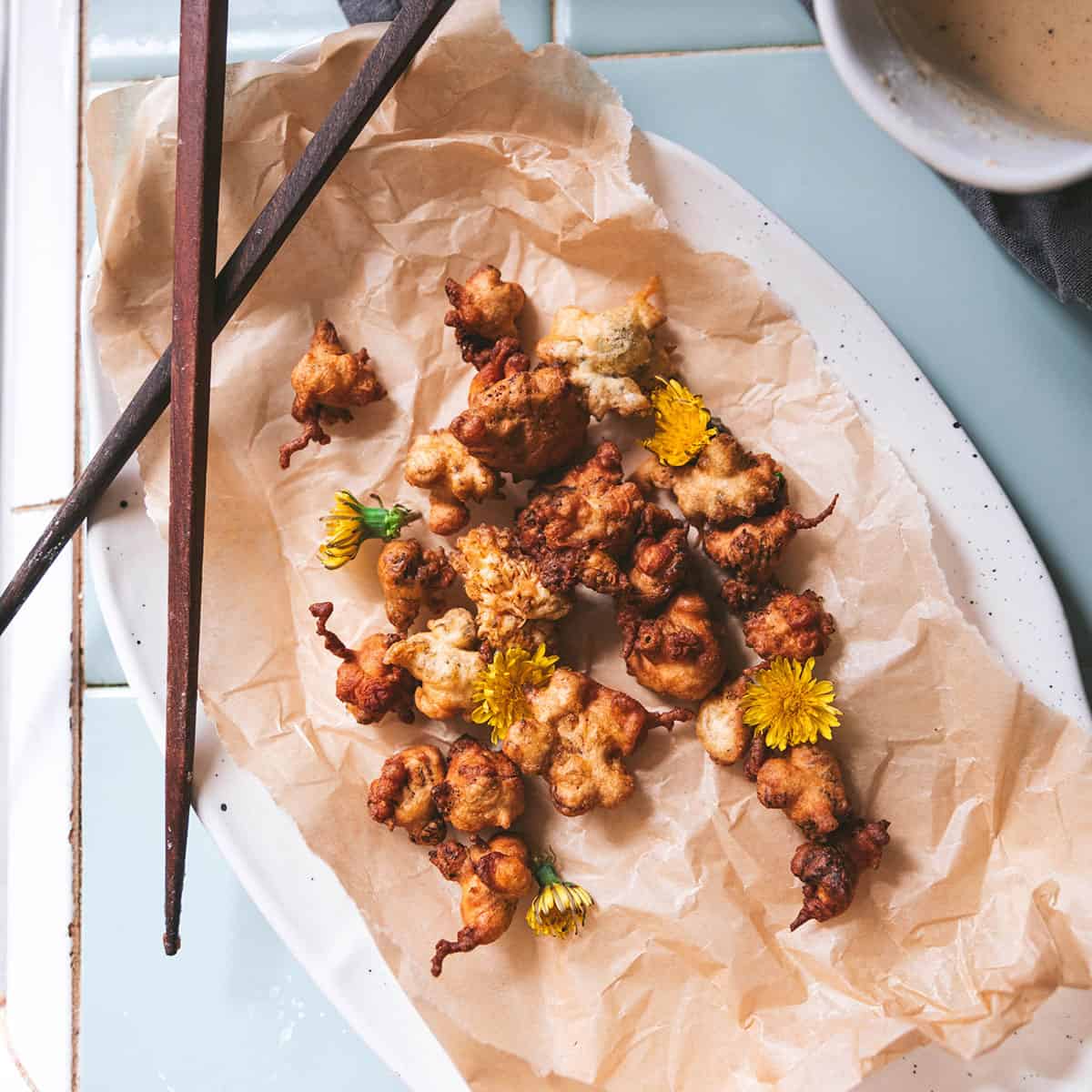 An oval plate with a natural napkin holding fried dandelion fritters, with chopsticks across the top of the plate. 