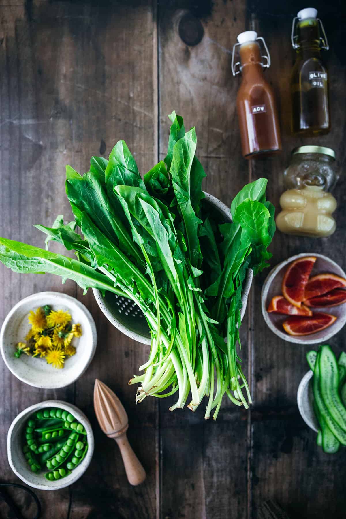 Fresh dandelion greens on a wood surface surrounded by small bowls of other salad ingredients, top view. 