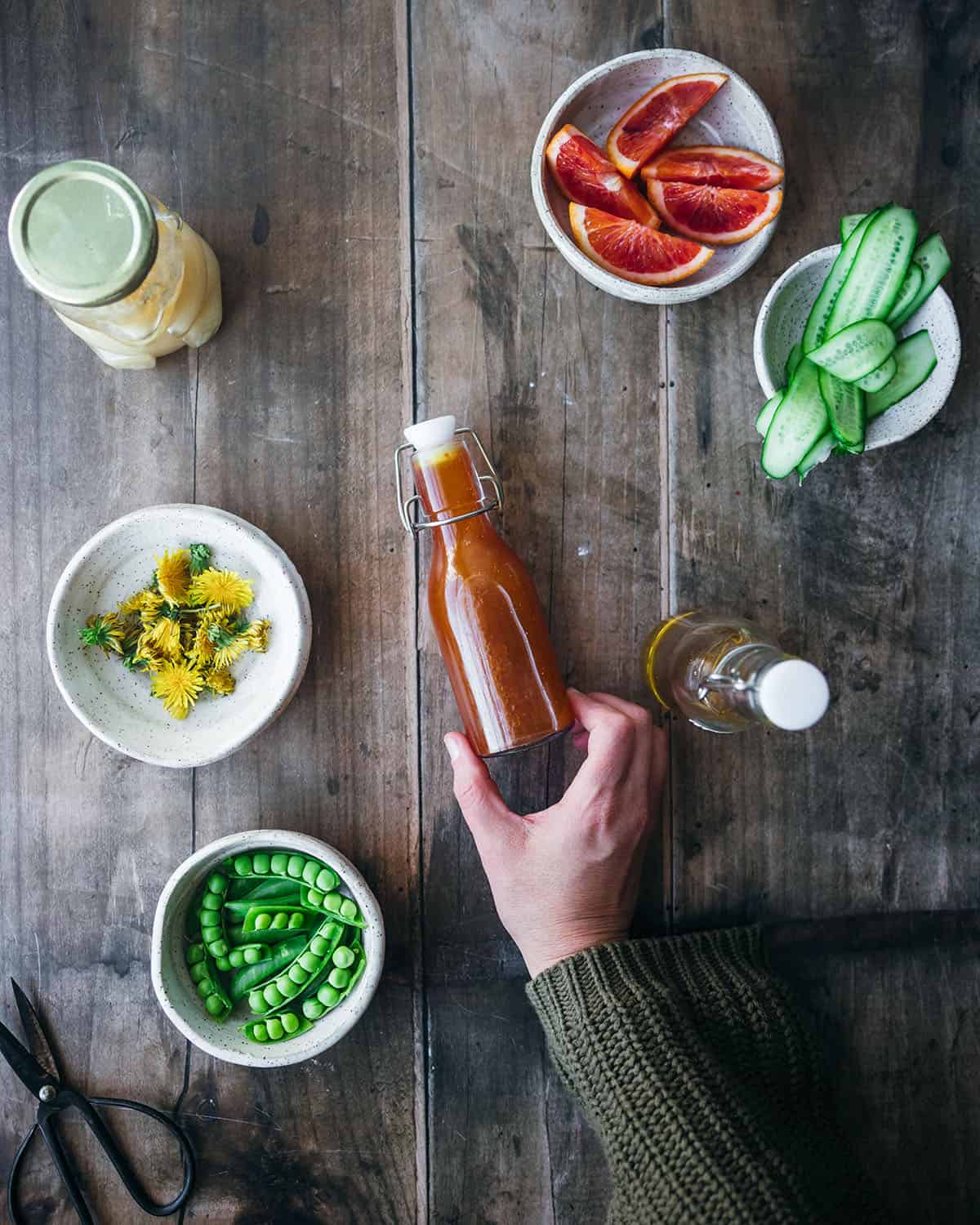 Dandelion salad dressing on a wood surface in a dressing jar with other dandelion salad ingredients surrounding. 