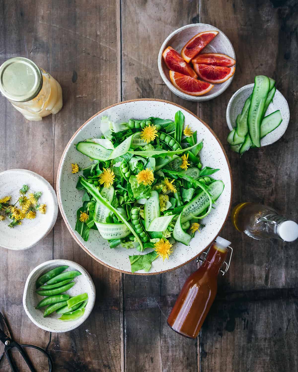 Dandelion salad in a white bowl, with other small bowls of ingredients surrounding, on a wood surface, top view. 