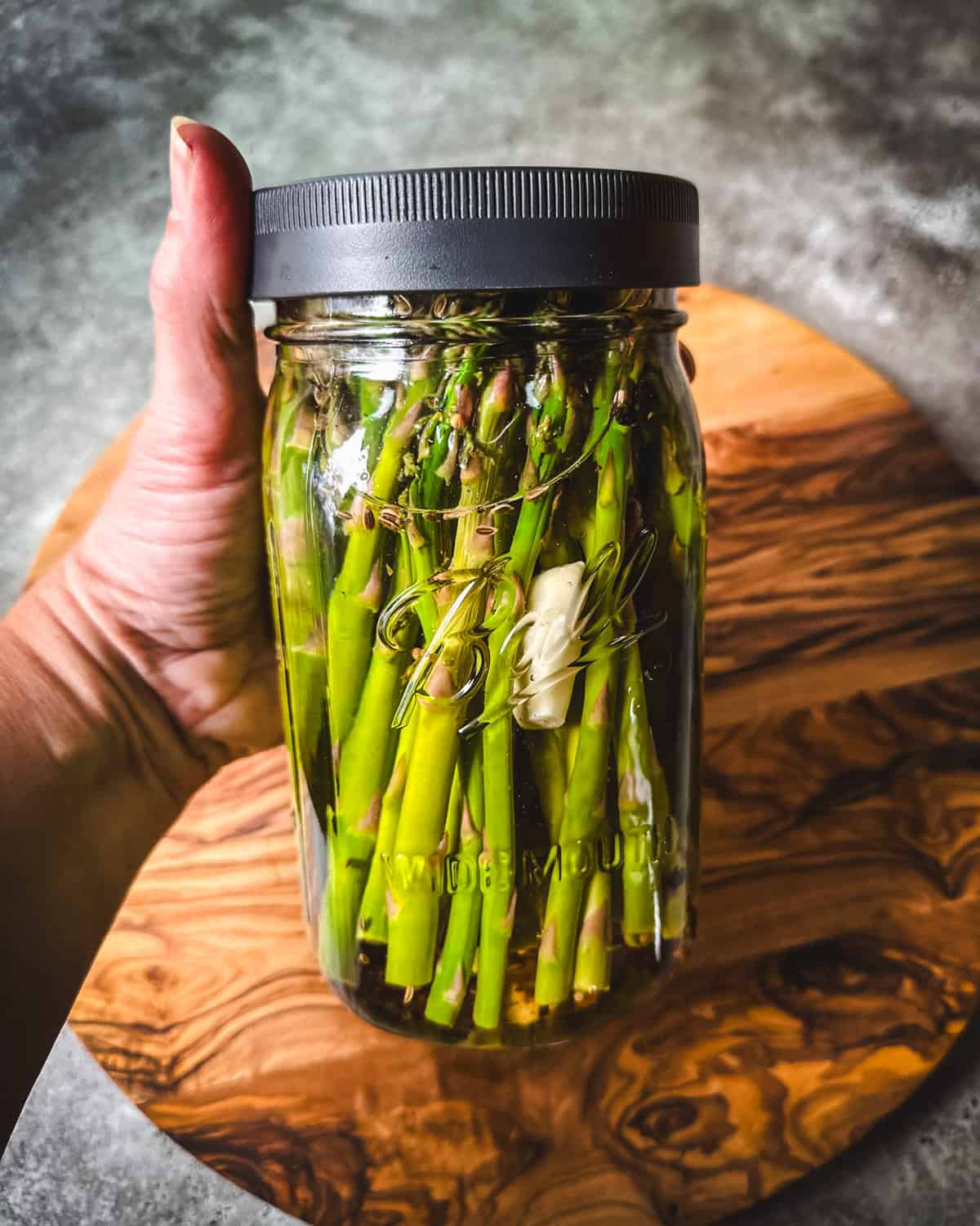 A jar of pickled asparagus with a lid on it, held out by a hand. 