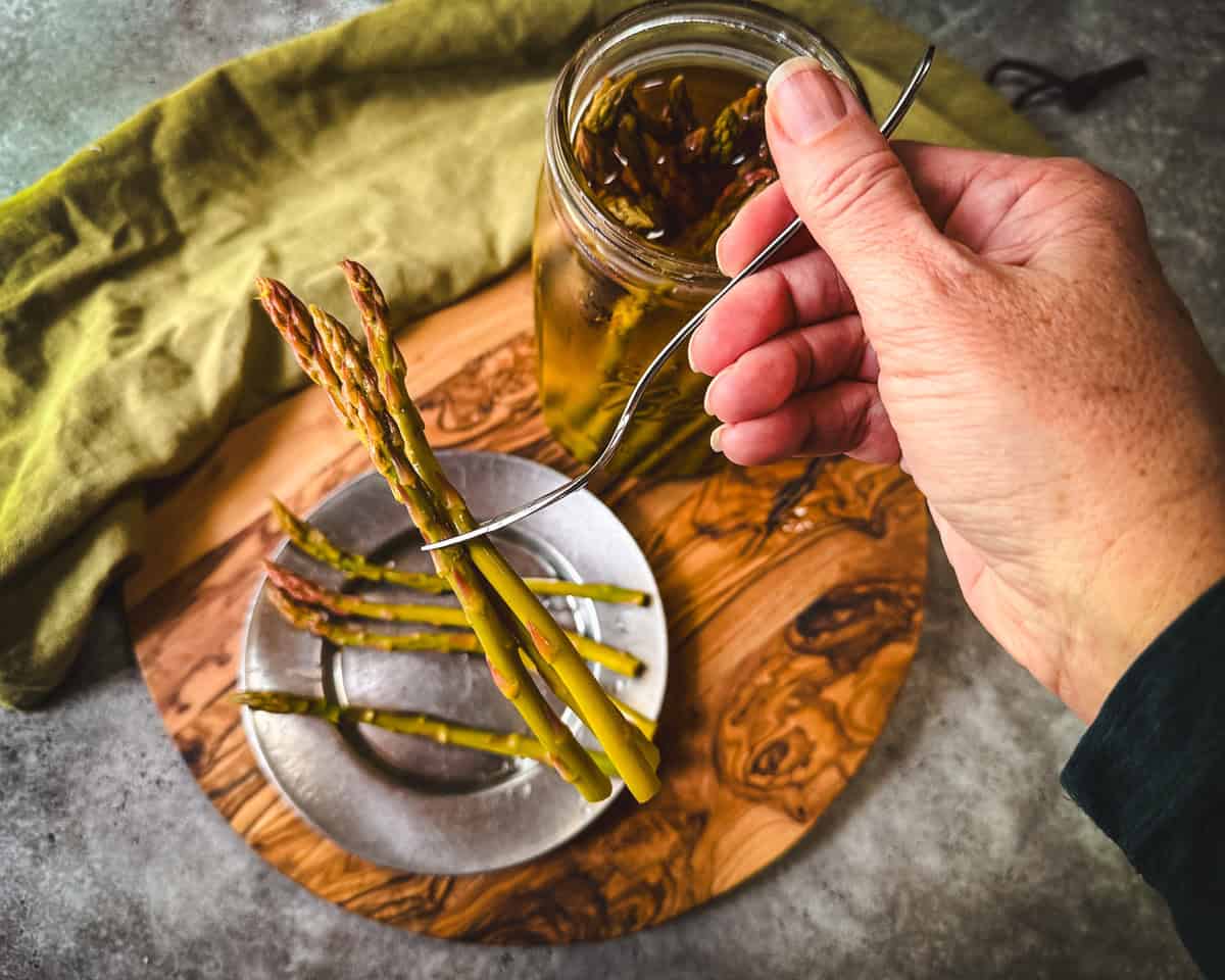 Pickled asparagus being lifted up with a fork over a metal plate and a wood cutting board surrounded by green fabric. 