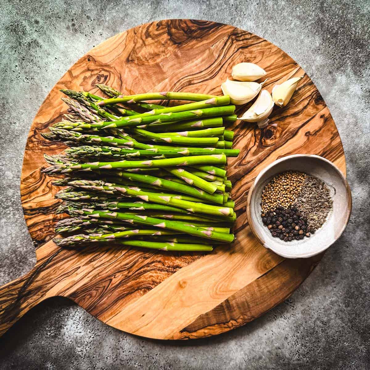 Pickled asparagus ingredients on a circular wood cutting board, top view. 