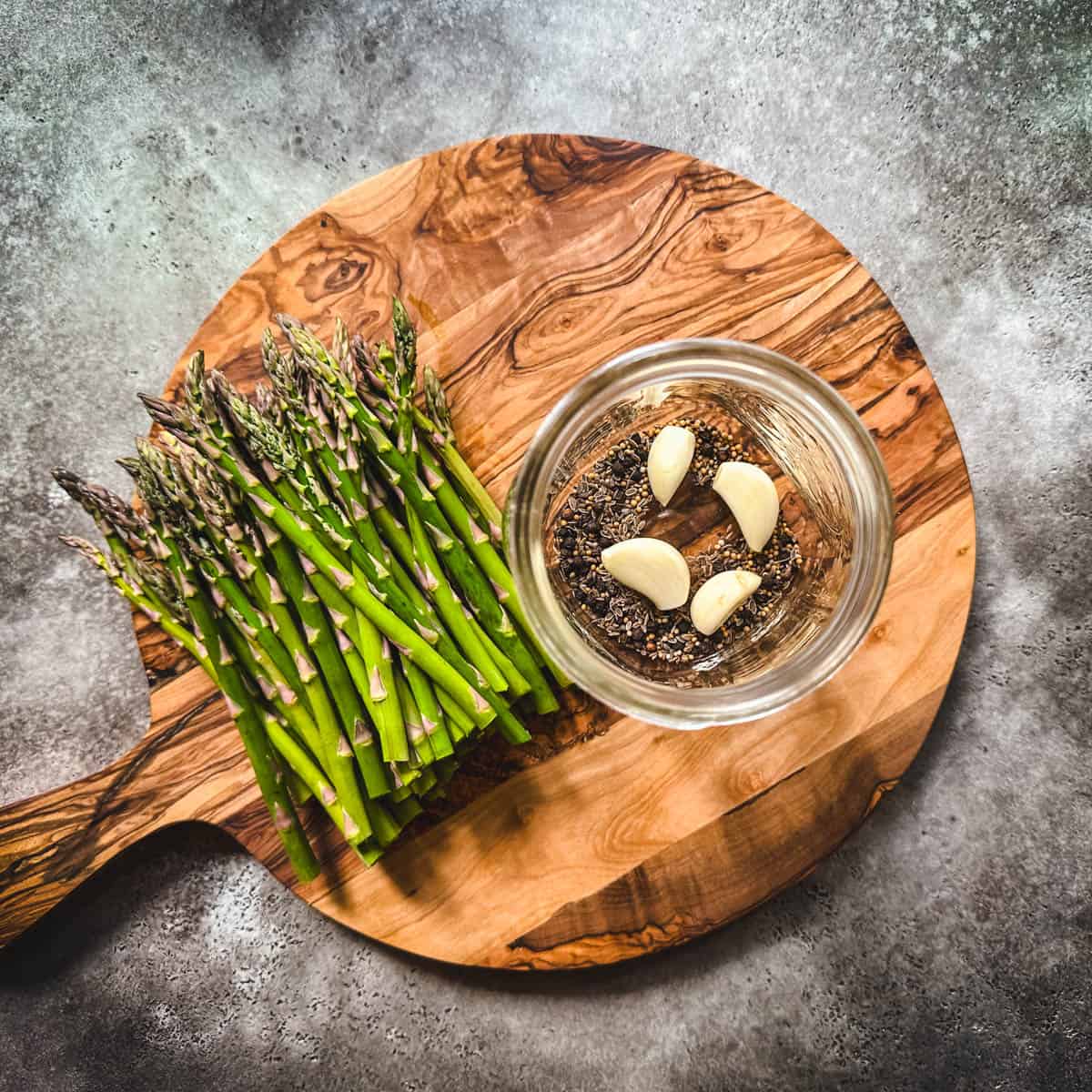 Asparagus trimmed next to a jar of pickling spices and garlic on a round wood cutting board, top view. 