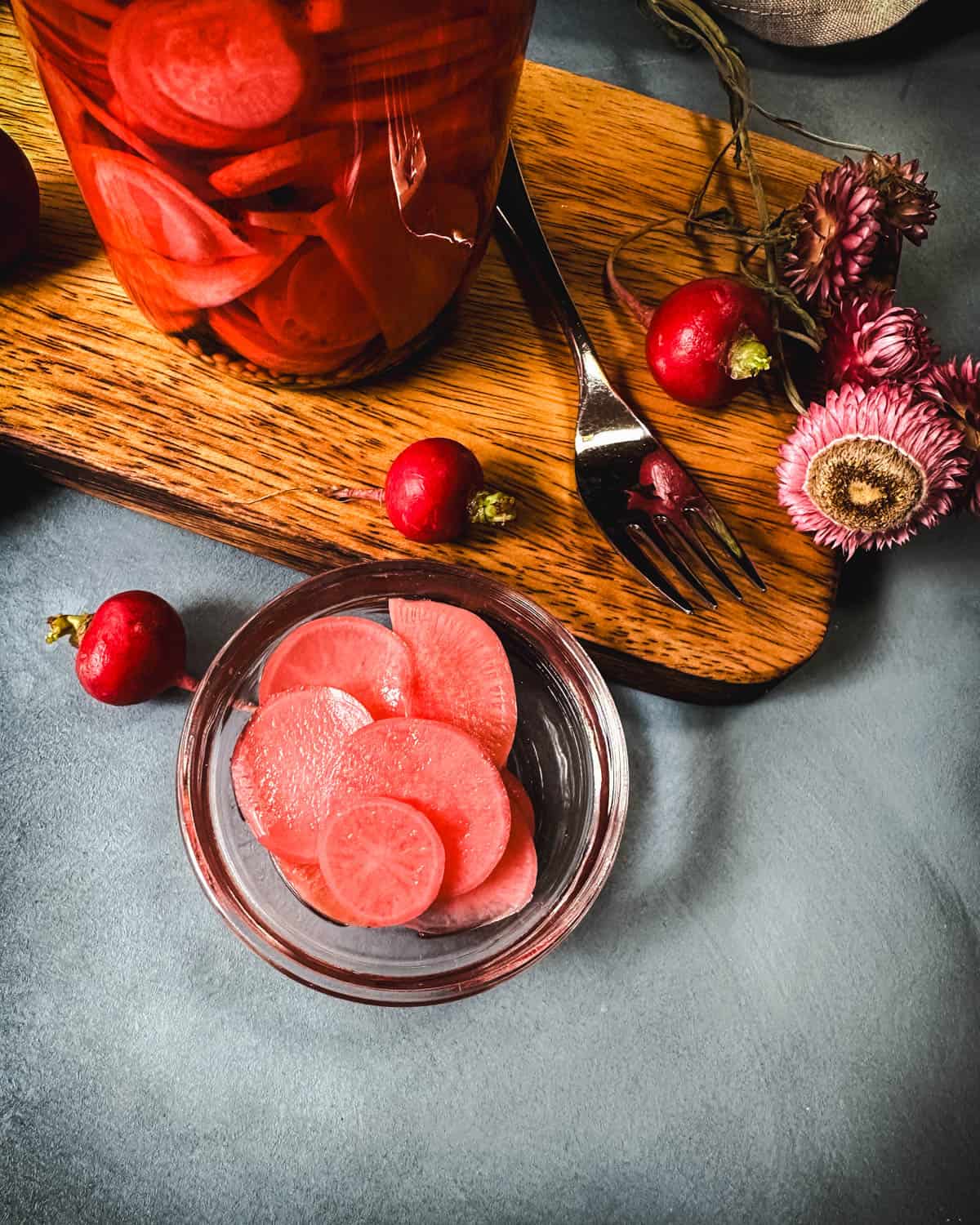 Pickled radishes in a jar, top view, with a wood cutting board to the side and fresh radishes and flowers surrounding.