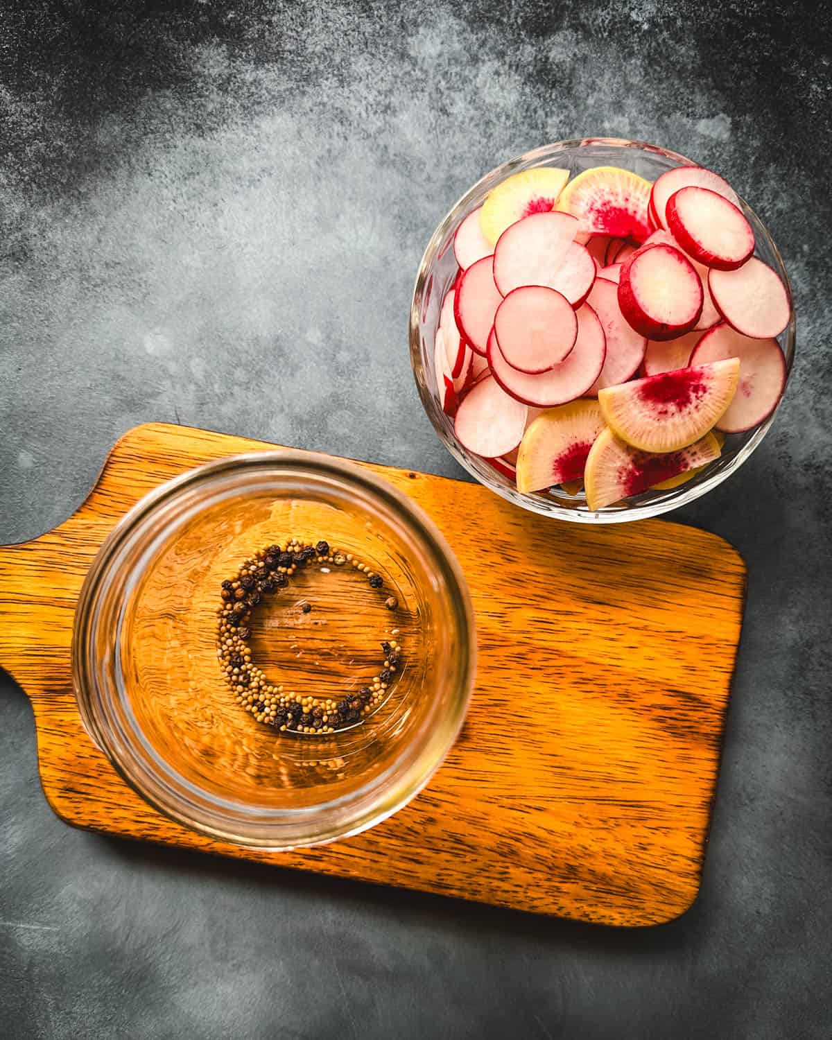 Sliced radishes in a bowl and pickling spices in a jar on a wood cutting board, top view. 