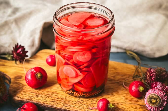 Pickled radishes in a mason jar on a wood cutting board with a white cloth in the background, with whole fresh radishes surrounding.