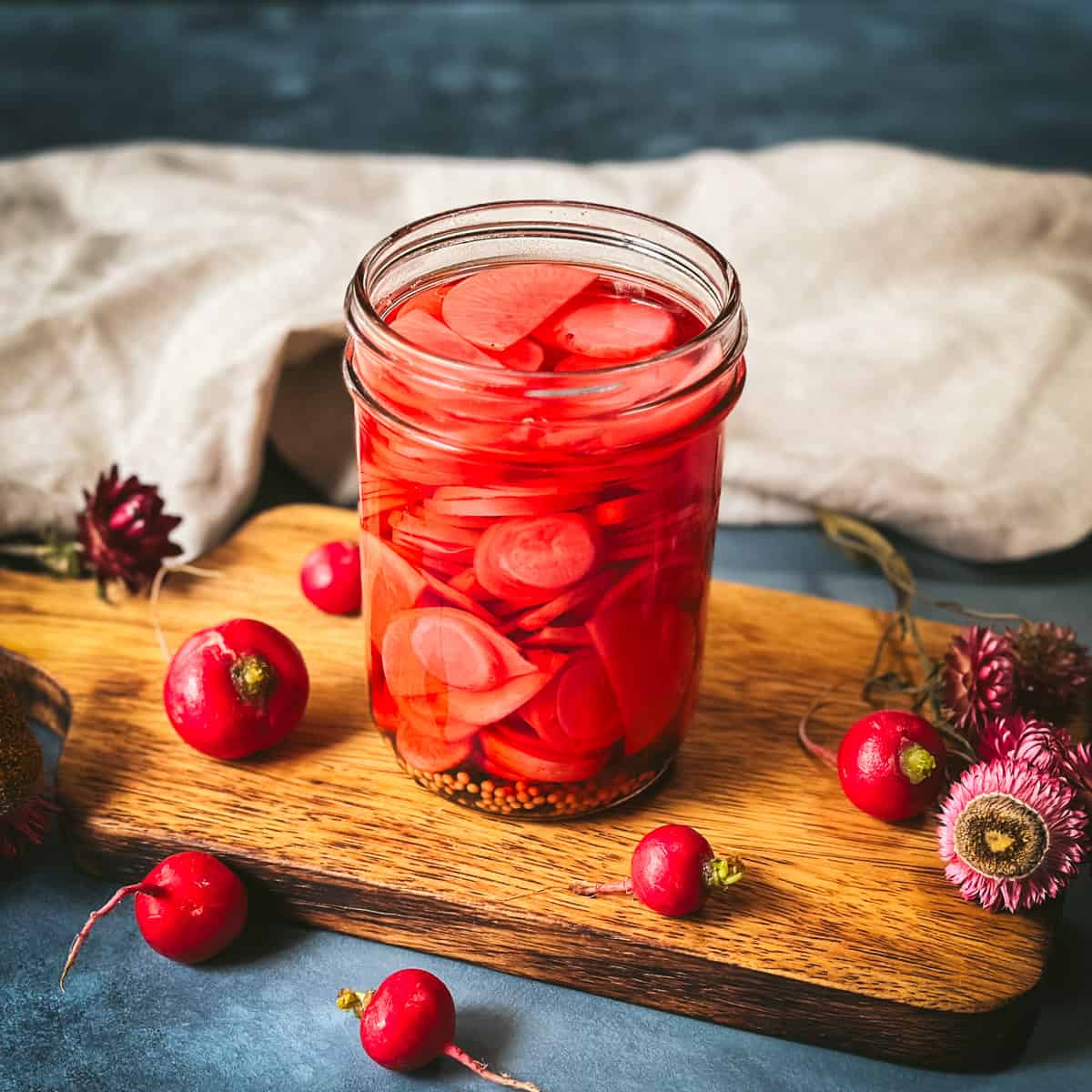 Pickled radishes in a mason jar on a wood cutting board with a white cloth in the background, with whole fresh radishes surrounding. 
