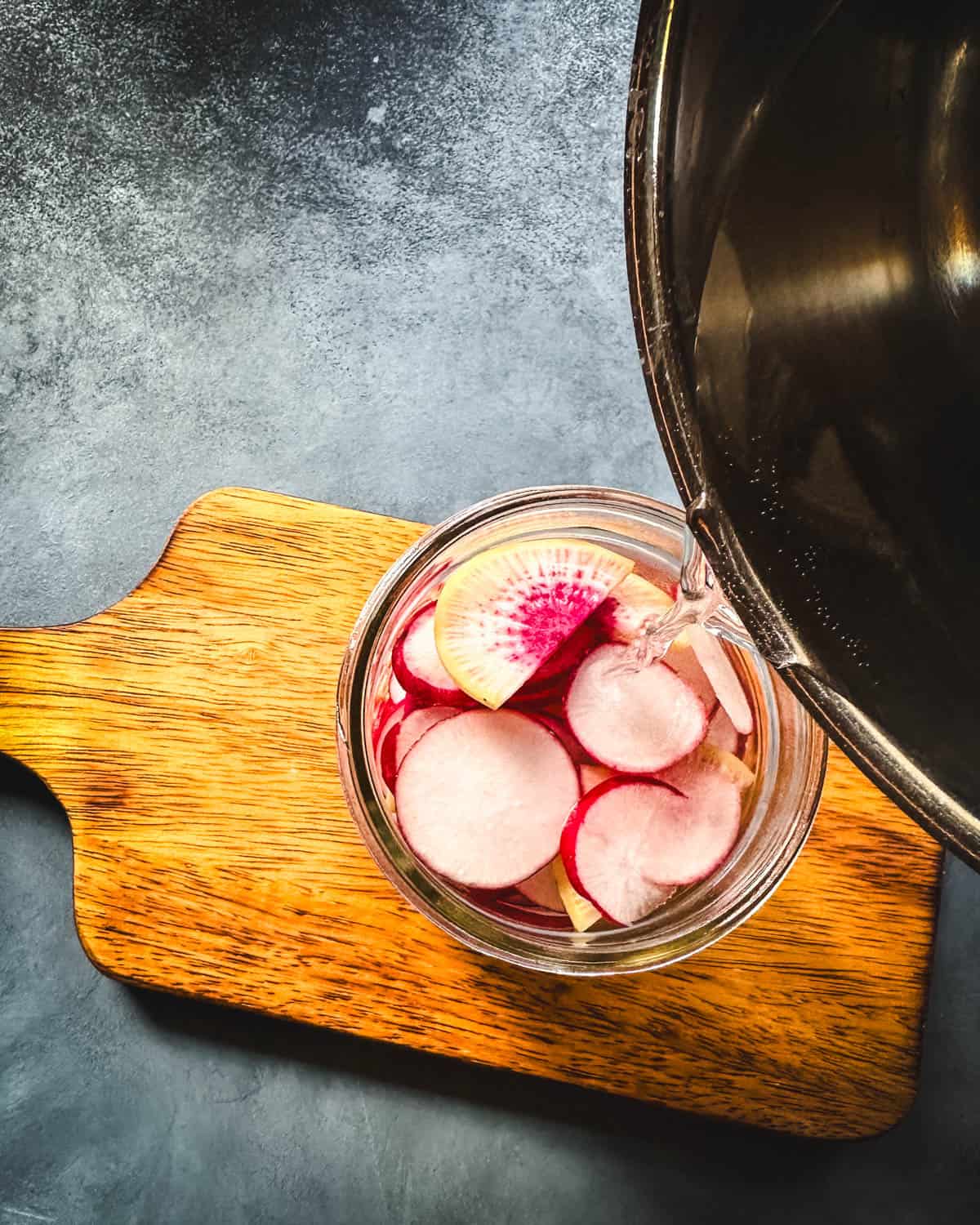 Brine pouring from a pan into a jar of sliced radishes on a wood cutting board, top view. 