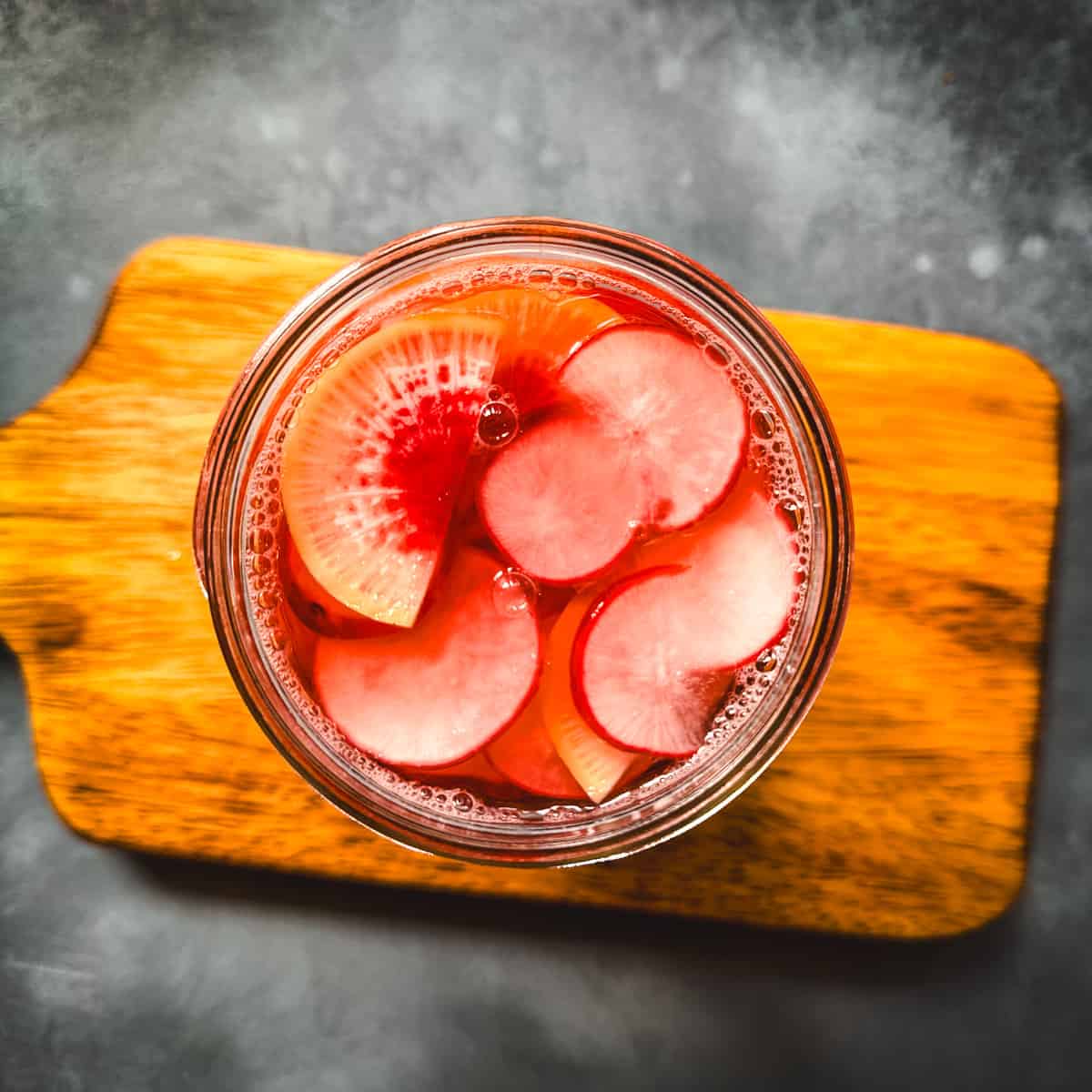 Pink pickled radishes in a jar on a wood cutting board, top view. 