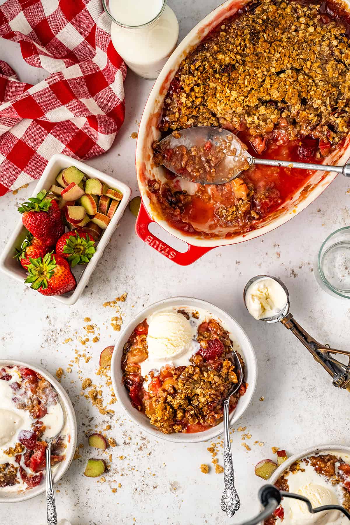 A red baking dish of strawberry rhubarb crisp with a serving spoon, on a white counter surrounded by a bowl of crisp with ice cream, an ice cream scoop, strawberries, and a red and white checkered towel. 
