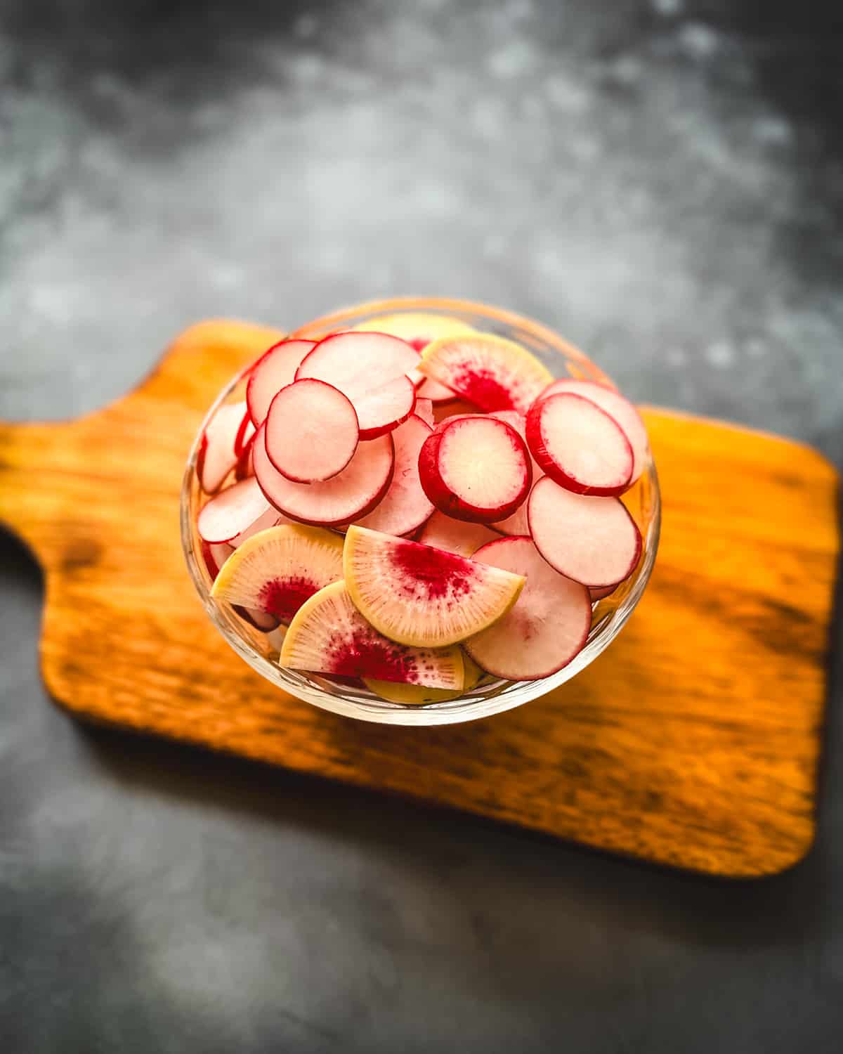 A bowl of thinly sliced radishes, on a wood cutting board. Top view. 