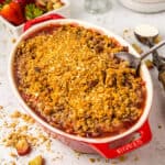 A red and white baking dish with rhubarb strawberry crisp with a serving spoon in it, surrounded by strawberries and oatmeal.