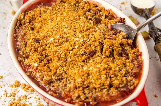 A red and white baking dish with rhubarb strawberry crisp with a serving spoon in it, surrounded by strawberries and oatmeal.