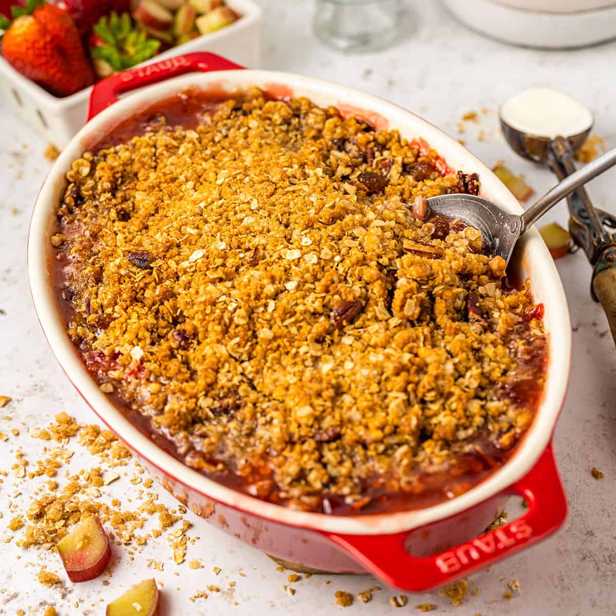 A red and white baking dish with rhubarb strawberry crisp with a serving spoon in it, surrounded by strawberries and oatmeal. 