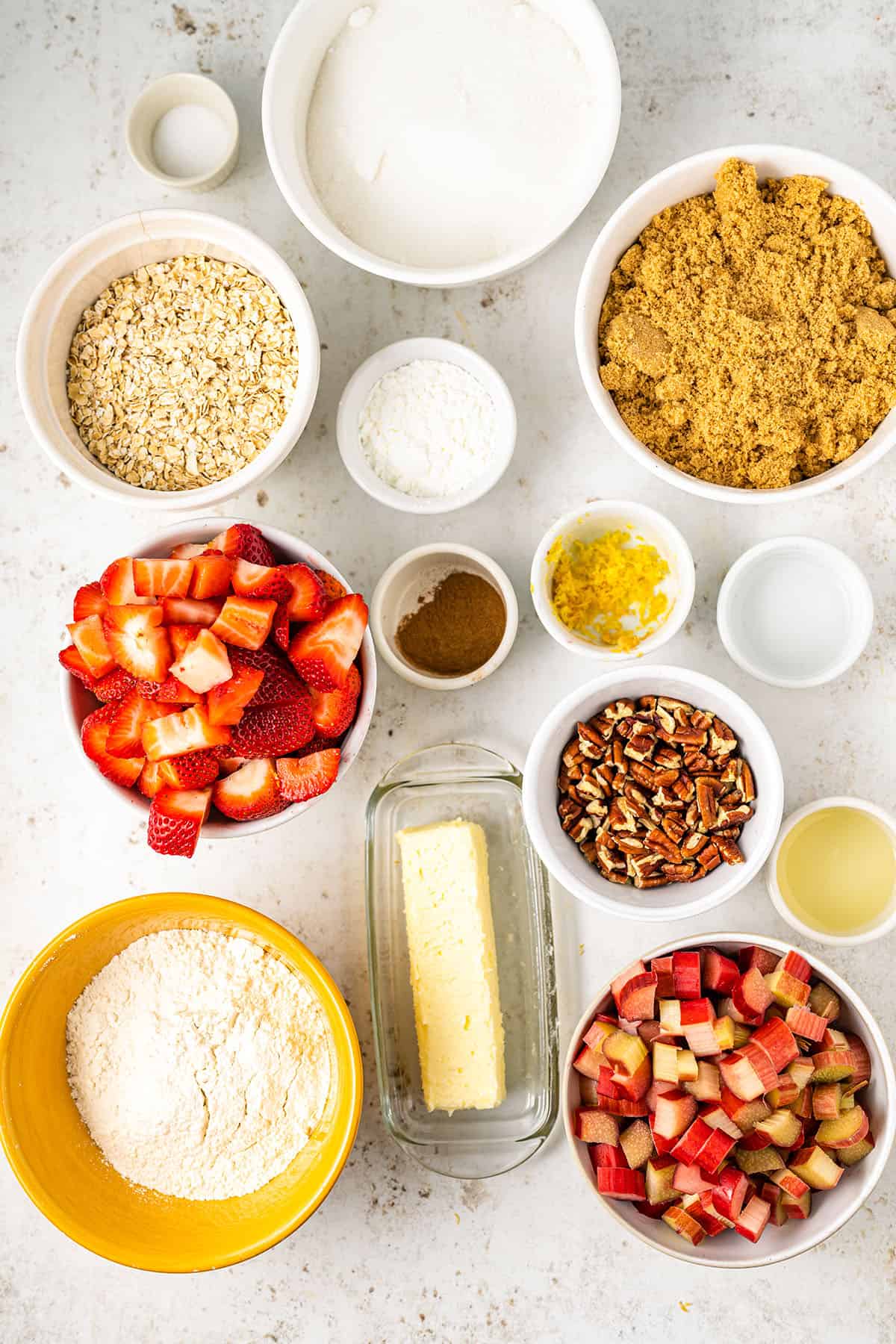 Ingredients for rhubarb strawberry crisp in bowls sitting on a white counter, top view. 