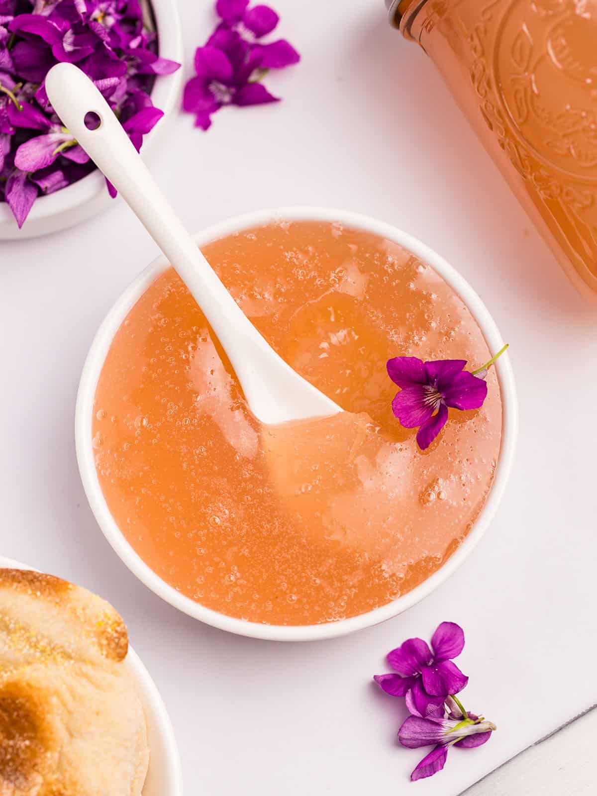 A white bowl of wild violet jelly with a white spoon, surrounded by fresh wild violet flowers. 