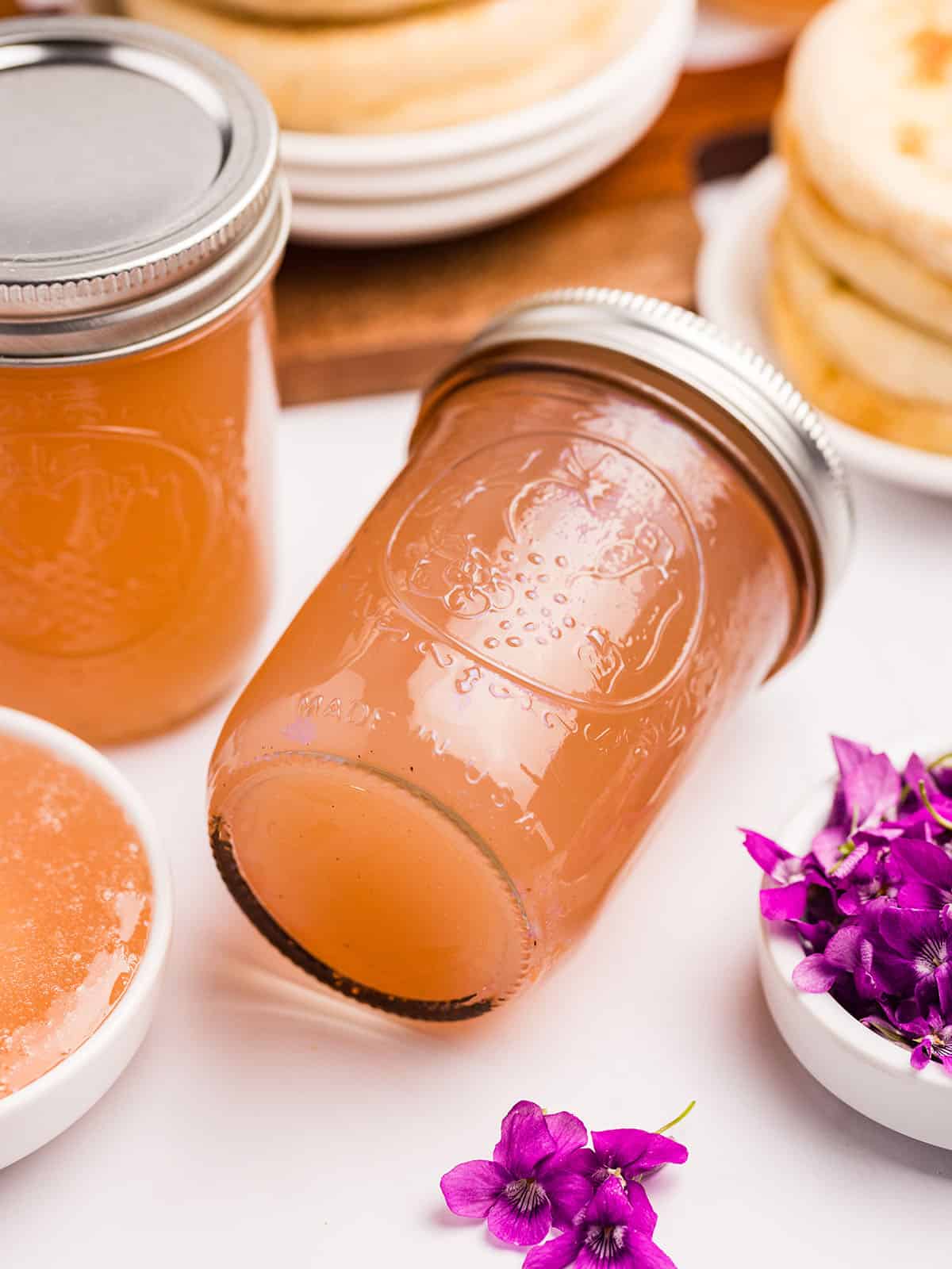 Jars of wild violet jelly, with one on it's side, surrounded by English muffins and wild violet flowers. 