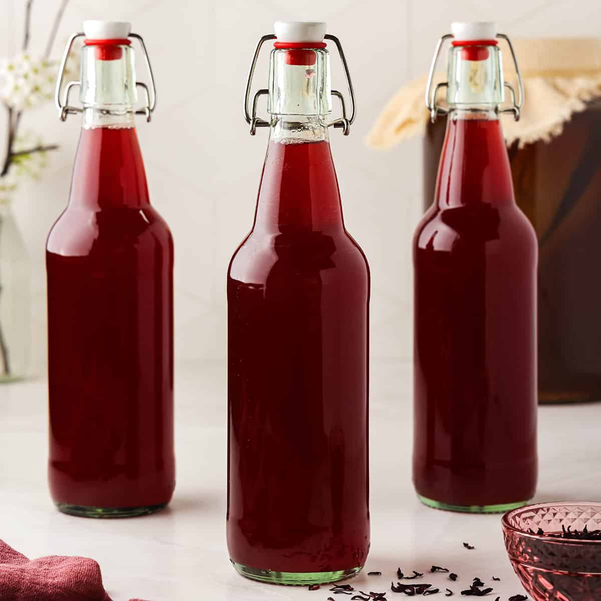 Three flip top bottles of deep pink hibiscus kombucha, on a white countertop, surrounded by dried hibiscus flowers.
